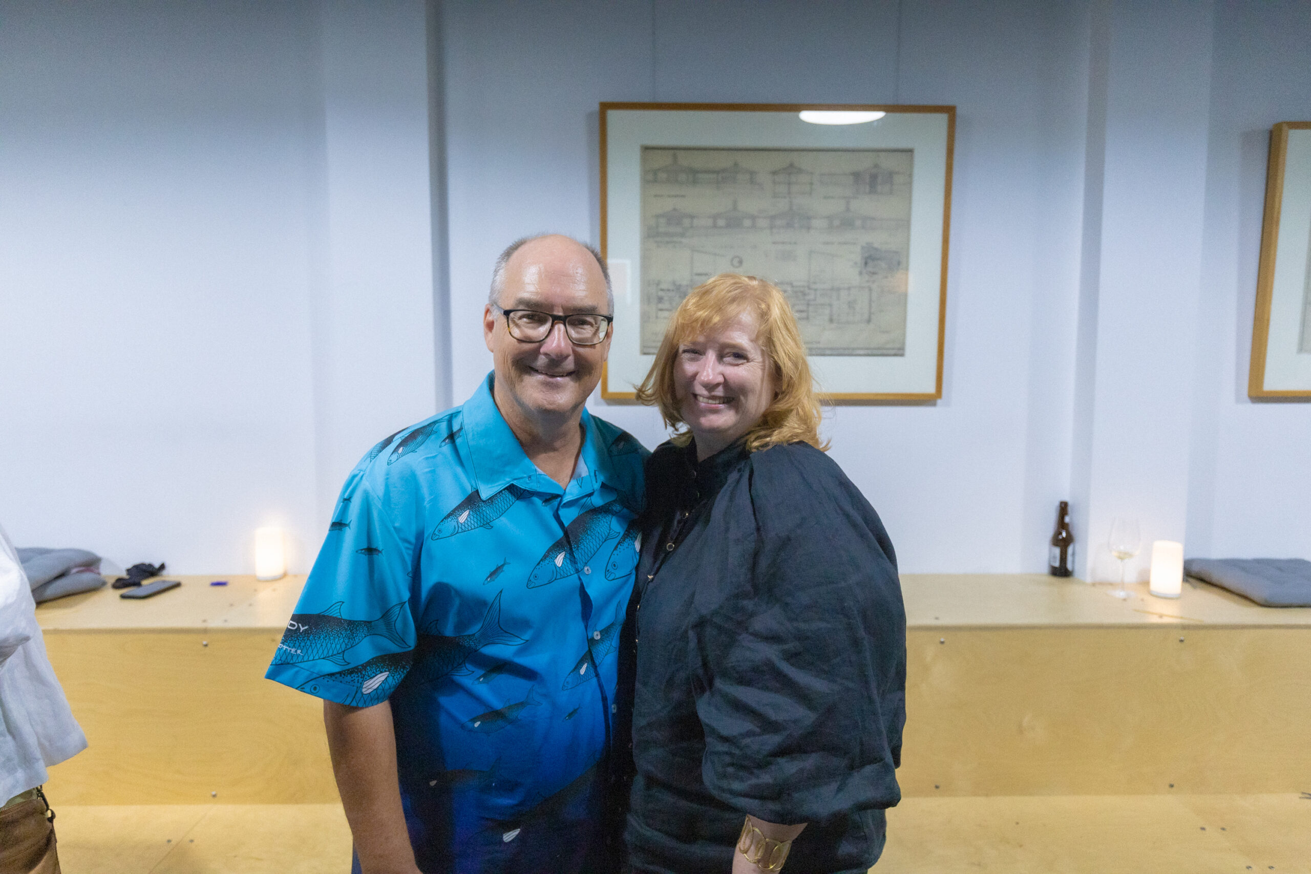Paul Trotter standing in a blue fishing shirt next to wife Mary, wearing a black blouse. The couple stand in front of a wooden staircase in the Fulton Trotter office.